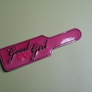 Pu&ograve; includere: Una paletta rosa con le parole "Good Girl" in corsivo e due cuori. La paletta ha un manico ed &egrave; bordata da cuciture nere. Lo sfondo &egrave; di colore verde chiaro.