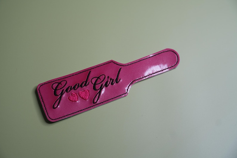 Pu&ograve; includere: Una paletta rosa con le parole "Good Girl" in corsivo e due cuori. La paletta ha un manico ed &egrave; bordata da cuciture nere. Lo sfondo &egrave; di colore verde chiaro.