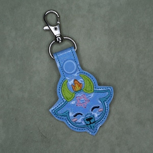 May include: A light blue embroidered keychain featuring a cartoon goat design with green horns and a flame. The keychain has a silver clasp and a blue snap closure. The goat has a smiling face and a pink floral design.