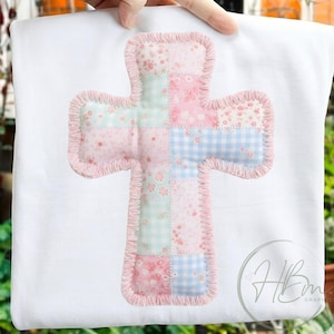以下が含まれることがあります： パッチワークの十字架のデザインが施された白い布地。十字架は、パステルカラーの花柄とギンガムチェックのパターンで構成され、ピンク色のフリルで縁取られています。デザインは手で持たれています。