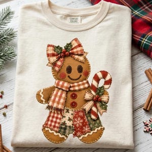 May include: An off-white t-shirt featuring a gingerbread person graphic. The gingerbread figure wears a red and white plaid bow, a matching scarf, and holds a candy cane. The figure has two red buttons on its chest and a patchwork-style skirt.