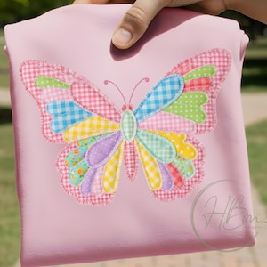 Op de afbeelding: Een lichtroze shirt met een grote vlinderapplicatie. De vleugels van de vlinder zijn gemaakt van verschillende stoffen met patronen, waaronder ruitjes, stippen en bloemenprints. Het lichaam van de vlinder is lichtgroen.