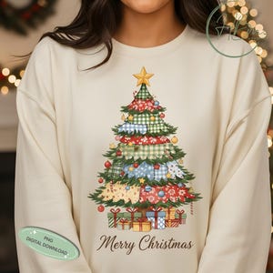 May include: Cream-colored sweatshirt featuring a Christmas tree design. The tree is decorated with various patterns and ornaments, topped with a gold star, and surrounded by wrapped gifts. The words "Merry Christmas" are written below the tree.