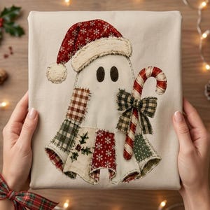 Op de afbeelding: Een beige sweatshirt met een centrale applicatie van een geest die een rode kerstmuts met witte sneeuwvlokken draagt. De geest is versierd met diverse stoffen patches in ruitjes, hulst en sneeuwvlokpatronen en houdt een rood-witte zuurstok vast.