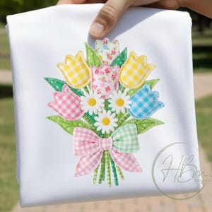 Peut inclure: Un tissu blanc orné d'une broderie florale. Le bouquet comprend des tulipes à carreaux jaunes, roses et bleues, des marguerites blanches et un nœud à carreaux roses et verts. Le motif comprend des feuilles et des tiges vertes.