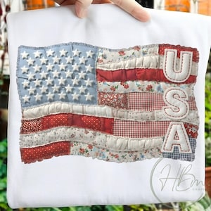 Puede incluir: Camiseta blanca con un diseño de bandera americana de patchwork. La bandera incluye tela azul, roja y blanca con las letras "USA" cosidas en el lado derecho. El diseño de la bandera tiene una estética vintage y desgastada.