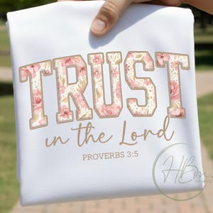 Könnte beinhalten: Weißes T-Shirt mit dem Schriftzug "TRUST in the Lord" in einem floralen Design. Der Text "PROVERBS 3:5" steht darunter. Das T-Shirt wird von einer Hand gehalten.