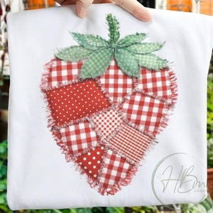 May include: White t-shirt featuring a large, patchwork strawberry design. The strawberry is composed of red and white gingham and polka dot fabrics, with green gingham leaves. The edges have a frayed, textured appearance.