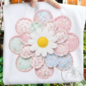 May include: White t-shirt with a large daisy applique. The daisy has a yellow centre and white petals. The flower's petals are surrounded by a ring of pink fabric. The flower's petals are made of various pastel fabrics.