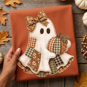 May include: A burnt orange t-shirt featuring a white ghost appliqué with dark brown eyes and a floral patterned bow. The ghost is decorated with various fabric patches in plaid and floral prints and holds a pumpkin with a green stem and leaf.