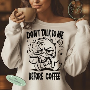 May include: White sweatshirt with the text "DON'T TALK TO ME BEFORE COFFEE" and a grumpy cartoon duck holding a coffee cup. The design is in black. The person in the image is holding a coffee cup.