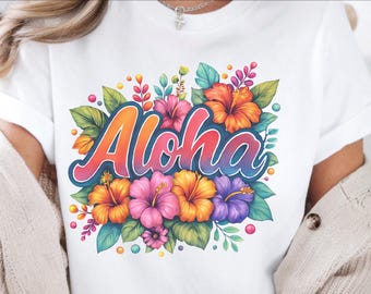 May include: White t-shirt featuring the word "Aloha" in a gradient orange and blue script, surrounded by colorful hibiscus flowers in shades of orange, pink, and purple. Green leaves and small orange and pink accents complete the tropical design.