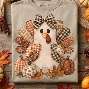 May include: A light beige t-shirt featuring a central applique of a turkey with a brown and white checkered bow. The turkey has patterned fabric feathers in shades of orange, brown, and cream, flanked by two fabric pumpkins on a textured base.