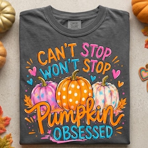 May include: A charcoal gray t-shirt featuring a graphic print with the text CAN'T STOP WON'T STOP PUMPKIN OBSESSED. The design includes three pumpkins in orange, pink, and white with polka dots and tie-dye patterns, surrounded by small hearts and leaves.