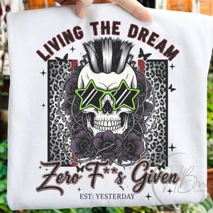 Puede incluir: Sudadera blanca con un diseño de calavera con gafas de sol en forma de estrella y cresta. El diseño incluye las frases "LIVING THE DREAM" y "Zero F's Given", con detalles florales y de estampado de leopardo. También está presente el texto "EST: YESTERDAY".