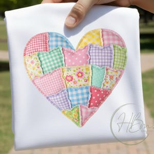 Op de afbeelding: Een wit stoffen item met een patchwork hart ontwerp. Het hart bestaat uit stofvierkanten in pastelkleuren, waaronder roze, blauw, groen en geel, met bloemen-, stippen- en ruitpatronen. Het item wordt door een hand vastgehouden.