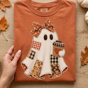 May include: A burnt orange t-shirt featuring a central applique of a white ghost with dark oval eyes. The ghost is adorned with a patterned bow and various fabric patches in plaid and floral prints, holding a coffee cup with a red heart.
