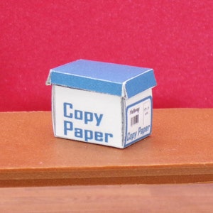 May include: A blue and white cardboard box with the words "Copy Paper" printed on the front. The box is sitting on a wooden table.