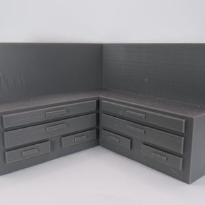 May include: A gray, corner-shaped miniature kitchen cabinet with multiple drawers. The cabinet is made of a solid material and has a textured surface. The drawers are rectangular with small, recessed handles.
