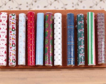 Könnte beinhalten: Zehn Rollen Geschenkpapier mit verschiedenen festlichen Mustern, darunter rot-weiß, grün-weiß und blau-weiß. Die Rollen sind in einer Reihe auf einer braunen Oberfläche angeordnet.