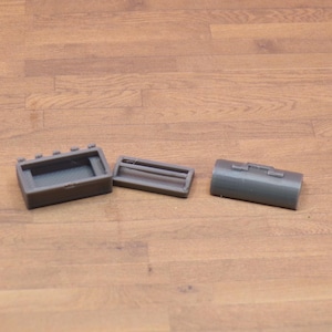 May include: Three gray 3D-printed objects are arranged on a wooden surface. The objects include a rectangular box with a grid, a rectangular frame, and a cylindrical container with a hinged lid. The items are likely components of a larger set.