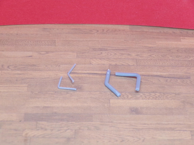 May include: Four light blue metal hooks are arranged on a wooden surface. Two hooks are L-shaped, and two are bent at an angle. The background features a red surface.