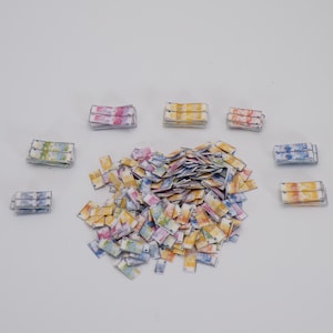 May include: A collection of small, colorful paper rectangles, resembling miniature stacks of money, are scattered across a white surface. The rectangles feature watercolor-like patterns in various hues, including pink, yellow, blue, and green.