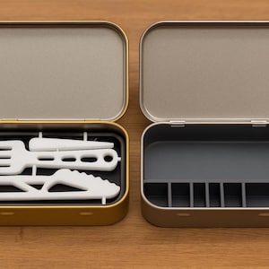 May include: Two open metal tins on a wooden surface. One tin is gold and contains white utensils: a fork, knife, and spoon. The other tin is silver and has a gray interior with dividers. The tins are rectangular with rounded corners.