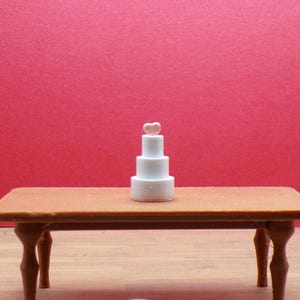 Puede incluir: Una pequeña tarta blanca de tres pisos con un adorno en forma de corazón se encuentra sobre una pequeña mesa marrón. La mesa tiene cuatro patas curvas. El fondo es de color rojo liso. La tarta es de temática nupcial o de celebración.