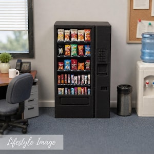 May include: A miniature black vending machine filled with various small snack bags and candy bars arranged on five shelves. The unit features a glass front, a keypad, a coin slot, and a dispensing tray, designed as a detailed scale model for display.