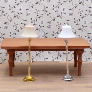 以下が含まれることがあります： 小さな木製のテーブルの上に、クリーム色と白の布製シェードが付いた2つのミニチュアランプが立っています。 ランプには装飾的なベースと細い茎があります。 テーブルは茶色で、4本の脚があります。 背景には花の壁紙があります。