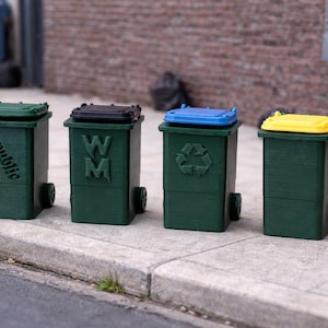 Puede incluir: Cuatro mini cubos de basura verdes con tapas de diferentes colores: verde, negro, azul y amarillo. Cada uno tiene ruedas y un logotipo o texto diferente: "Republic", "WM", un símbolo de reciclaje y ninguno. Están alineados en una acera de hormigón.