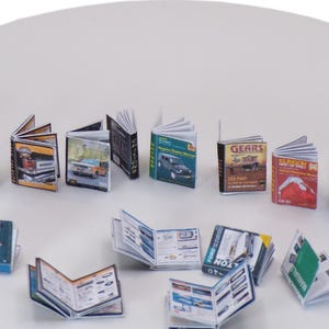 Puede incluir: Una colección de libros en miniatura, algunos con cubiertas de colores y otros con cubiertas en blanco y negro. Los libros están dispuestos en una fila sobre una superficie blanca.