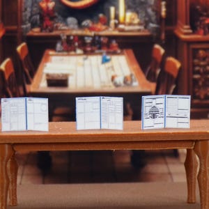 Puede incluir: Tres libros en miniatura, blancos, encuadernados en papel con cubiertas azules y texto negro se muestran en una mesa de madera marrón. Los libros están dispuestos en fila, con las cubiertas mirando al espectador.