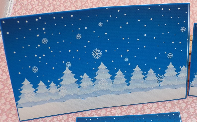 K&ouml;nnte beinhalten: Eine blaue Karte mit einer Winterszene aus wei&szlig;en B&auml;umen und Schneeflocken vor blauem Hintergrund.