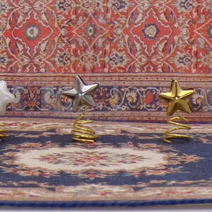 May include: Three miniature Christmas tree toppers, each with a star-shaped design. The stars are white, silver, and gold. The toppers are on a spring base and are sitting on a blue and gold patterned rug.