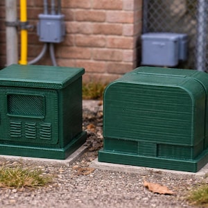 May include: Two dark green utility boxes, one rectangular and one rounded, sit on concrete pads. The rectangular box has a door with a window and vents. The rounded box has a curved top. Both are outdoors near a brick wall and a chain-link fence.
