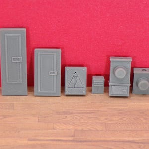 May include: A collection of gray miniature electrical boxes and panels, including a tall door-like panel, a smaller door-like panel, a box with a lightning bolt symbol, and other box-shaped objects. The items are on a wooden surface with a red background.