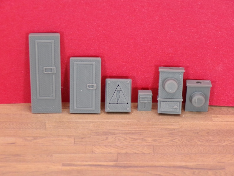 May include: A collection of gray miniature electrical boxes and panels, including a tall door-like panel, a smaller door-like panel, a box with a lightning bolt symbol, and other box-shaped objects. The items are on a wooden surface with a red background.
