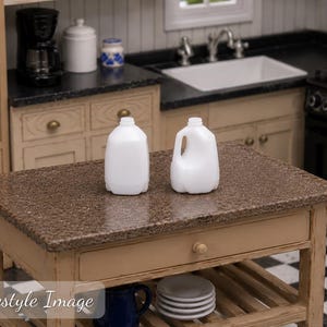 Peut inclure: Deux bidons de lait miniatures en plastique blanc avec poignées et ouvertures supérieures. Ces petits récipients sont conçus pour des cuisines de maison de poupée, avec une finition opaque lisse et une forme classique de bidon, posés sur un îlot.
