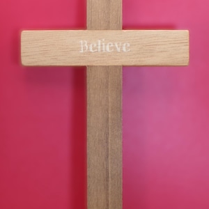 May include: A wooden cross with the word "Believe" painted in white on the horizontal beam. The cross is standing on a wooden surface in front of a pink background.