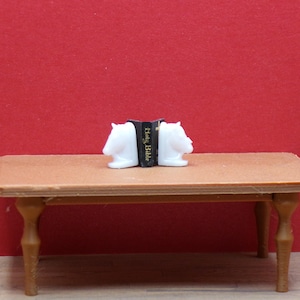 May include: Miniature scene featuring a small brown table with four legs. On the table sits a book held between two white horse head bookends. The book is black with gold lettering that reads "Holy Bible". The background is a solid red.