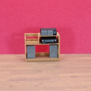 Op de afbeelding: Een miniatuur beige entertainment center met twee luidsprekers, een stereoreceiver en een cassettespeler. De luidsprekers en componenten zijn grijs en zwart. De achtergrond is effen roze.