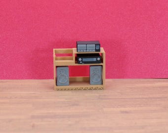 Può includere: Un mobile da intrattenimento beige in miniatura con due altoparlanti, un ricevitore stereo e un lettore di cassette. Gli altoparlanti e i componenti sono grigi e neri. Lo sfondo è di colore rosa.