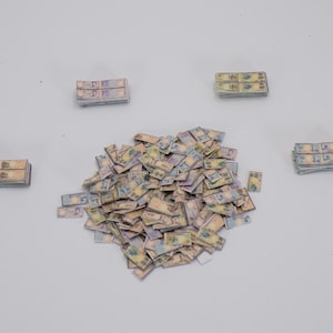 May include: A collection of miniature paper currency, with stacks of bills scattered around a larger pile. The bills have a mix of yellow, blue, and purple hues, suggesting a variety of denominations.