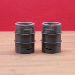 May include: Two dark gray, cylindrical objects with a series of stacked rings and a slot near the base. The objects have a textured appearance and are set against a red background on a wooden surface.
