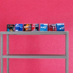 Può includere: Scatole di ricambi auto in miniatura, tra cui marchi come Autolite e Champion, disposte su una mensola grigia contro uno sfondo rosa. Le scatole sono di vari colori, con testo e grafica. La mensola è a due livelli.