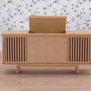 May include: A miniature wooden cabinet with a light brown finish. The cabinet has a rectangular shape with vertical slats on the sides and a textured center panel. It sits on short, angled legs, and has a slot on top.