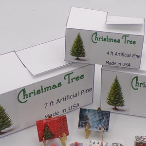 Può includere: Tre scatole di cartone bianco con testo verde e immagini di alberi di Natale. Le scatole sono etichettate "Christmas Tree" e indicano la dimensione dell'albero in piedi. Le scatole sono per alberi di pino artificiali da 4 piedi, 5 piedi e 7 piedi. Tutte le scatole indicano "Made in USA".
