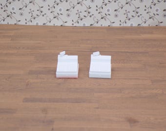 May include: Two white miniature cash registers with a small display screen on top. The registers are on a wooden surface with a floral wallpaper background. The registers have a grid-like pattern on the front.